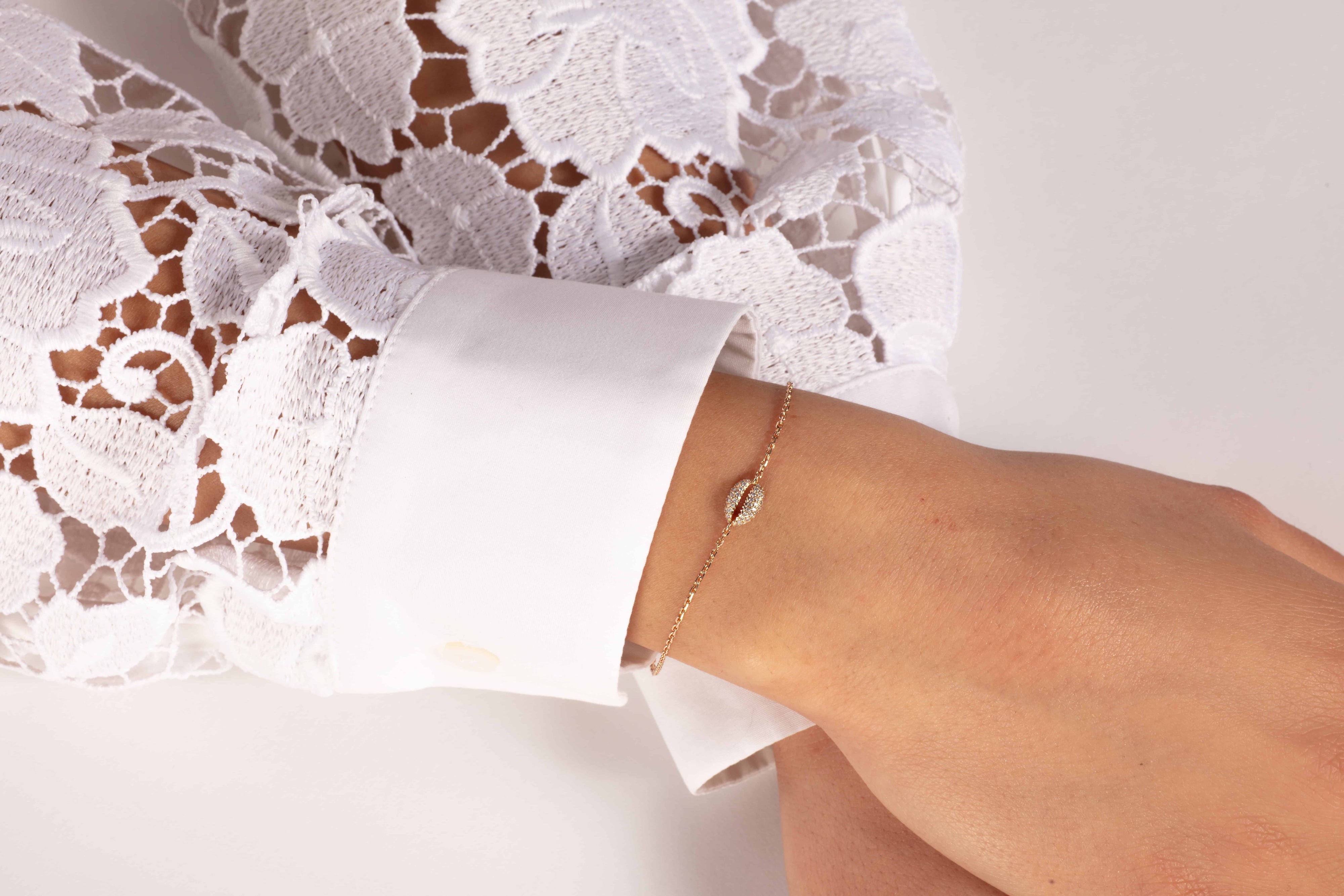 Eine Person trägt ein weißes Spitzentop, legt ihre Hand sanft auf eine Oberfläche und präsentiert ihr Handgelenk, das mit RHEA ROSÉGOLD von Atelier Sofia geschmückt ist, einem Armband aus 18 Karat Roségold mit einem eleganten blattförmigen Anhänger.