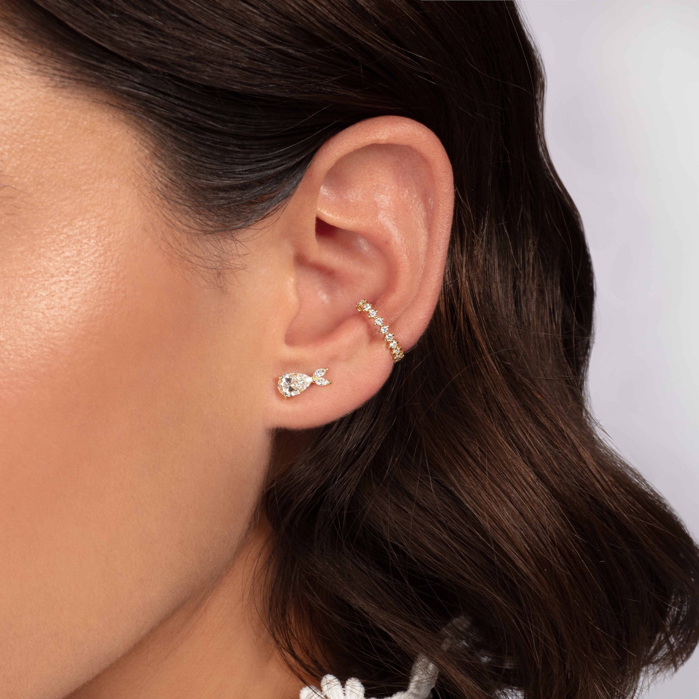 Eine Nahaufnahme zeigt eine Person mit kurzen braunen Haaren, die 18 K Weißgoldschmuck von Atelier Sofia trägt: einen goldenen Ear Cuff und einen fischförmigen Ohrstecker von Dione mit einem kleinen im Labor gezüchteten Diamanten. Der unscharfe Hintergrund betont die atemberaubenden DIONE WEIßGOLD-Schmuckstücke am Ohr der Person.